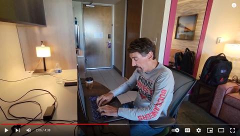 Screenshot from YouTube video of Bruce Wawrzyniak at desk in hotel on laptop Screenshot from YouTube video of Bruce Wawrzyniak at desk in hotel on laptop