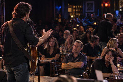 A male musician is seen from the back onstage with audience members looking bored and turned off as he speaks