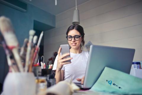 Girl seated at laptop but holding and looking at smartphone