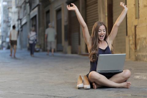 Girl sitting in street with laptop on knees while raising arms excitedly Girl sitting in street with laptop on knees while raising arms excitedly