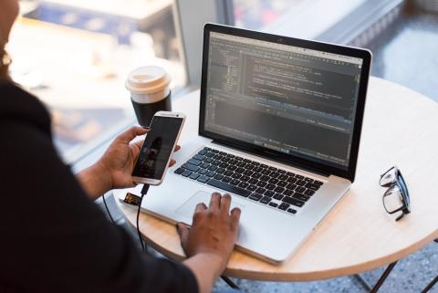 A female hand holds a smartphone with the other hand on a laptop keyboard with the screen displaying data A female hand holds a smartphone with the other hand on a laptop keyboard with the screen displaying data