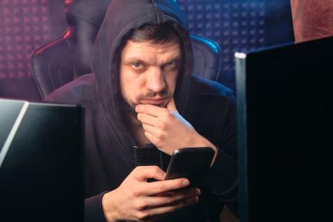 Male with hood up looking at cellphone while next to computer screen Male with hood up looking at cellphone while next to computer screen