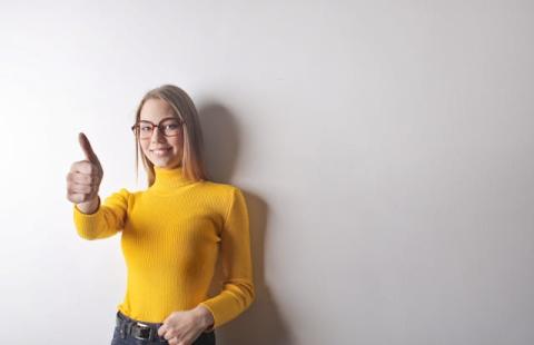 Girl indoors against plain wall wearing yellow shirt and glasses giving thumbs up Girl indoors against plain wall wearing yellow shirt and glasses giving thumbs up