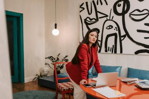 A girl in a red blazer leans at a table with a laptop and phone on it and abstract art on the wall A girl in a red blazer leans at a table with a laptop and phone on it and abstract art on the wall